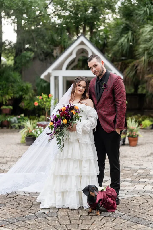 Alexandra's long veil trails beside Jason as they stand together at Riverhouse Events with their bouquets and dog.