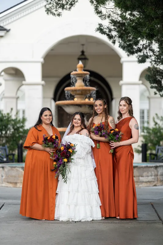 Alexandra in white stands with three bridesmaids in burnt orange dresses before Riverhouse Events' arched fountain, all holding bouquets.
