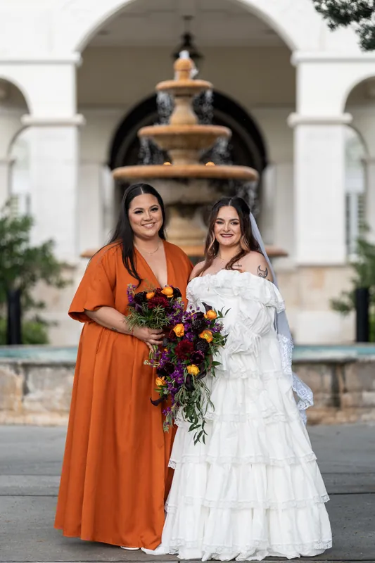 Alexandra and a bridesmaid in coral embrace before the tiered fountain at Riverhouse Events, holding their cascading floral arrangements.