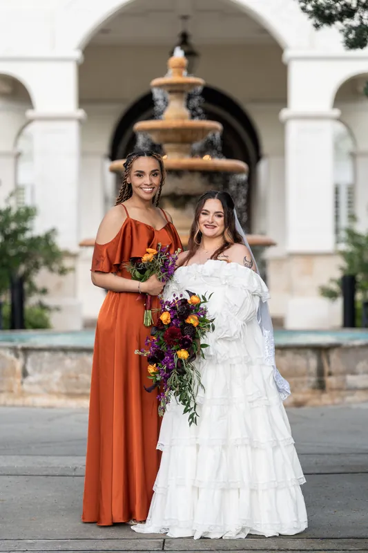 Two bridesmaids in matching burnt orange dresses flank Alexandra in her tiered white gown at Riverhouse Events' fountain courtyard.