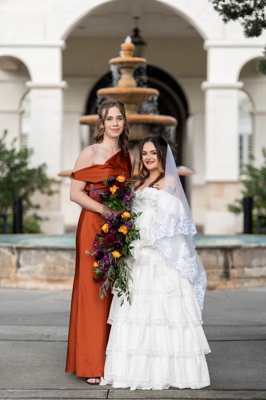 Alexandra in an orange off-shoulder gown and a bridesmaid in white stand before a fountain at Riverhouse Events, holding a cascading floral bouquet.