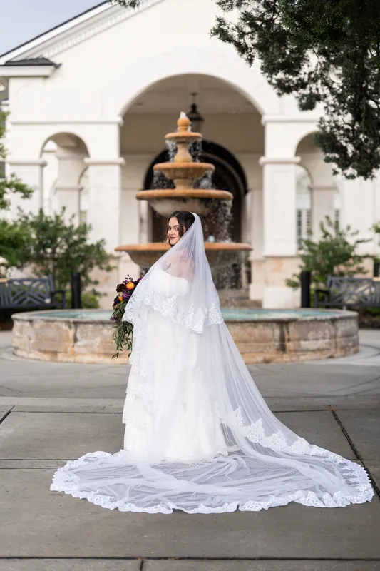 Alexandra in a layered white wedding dress with veil stands alone at Riverhouse Events, train spread across the stone courtyard.