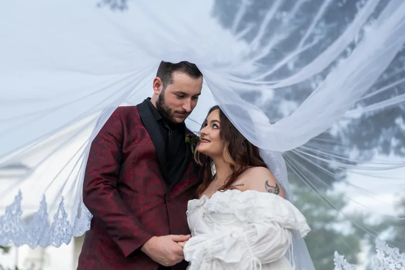 Jason and Alexandra embrace under flowing white fabric, gazing at each other with mountainous scenery behind them.