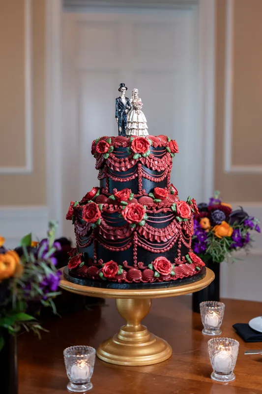A tiered wedding cake decorated with red roses and gold beading sits on a brass stand at Riverhouse Events, surrounded by purple flowers.