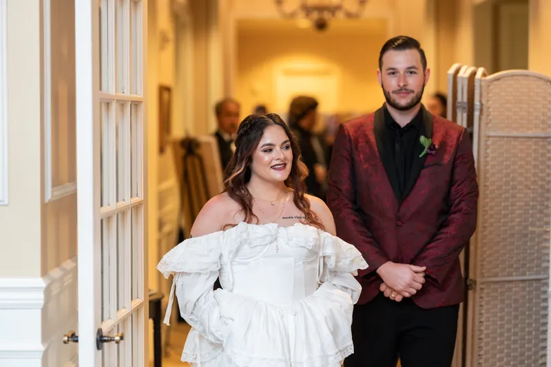 Alexandra and Jason stand together at Riverhouse Events, she in an off-shoulder white gown and he in a burgundy jacket, both smiling warmly.