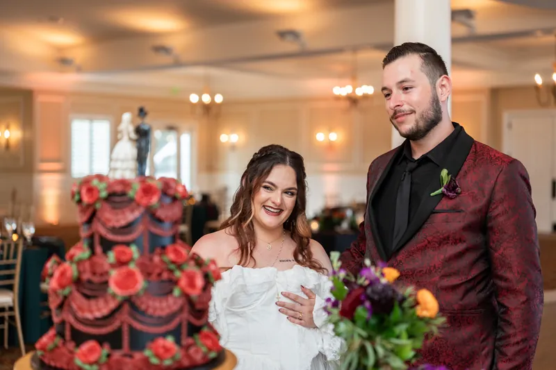 Alexandra beams beside Jason at Riverhouse Events, with a tiered red rose cake and floral arrangements visible behind them.