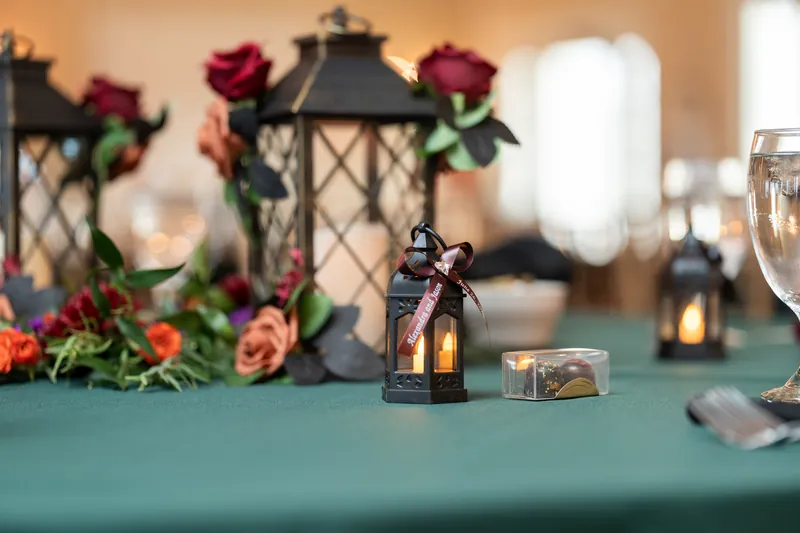 A black lantern centerpiece with lit candles sits on a teal-draped table surrounded by red and coral roses at Riverhouse Events.