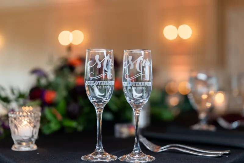 Two champagne flutes engraved with initials rest on a black tablecloth, glowing amber lights and greenery blurred softly behind.