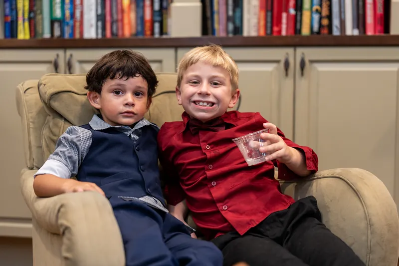 Two young boys sit together on a cream sofa, one in navy and one in burgundy, grinning at the camera with bookshelves behind them.