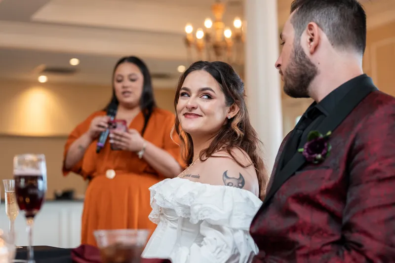 Jason listens attentively as Alexandra speaks during toasts, with a woman in orange holding a microphone nearby.