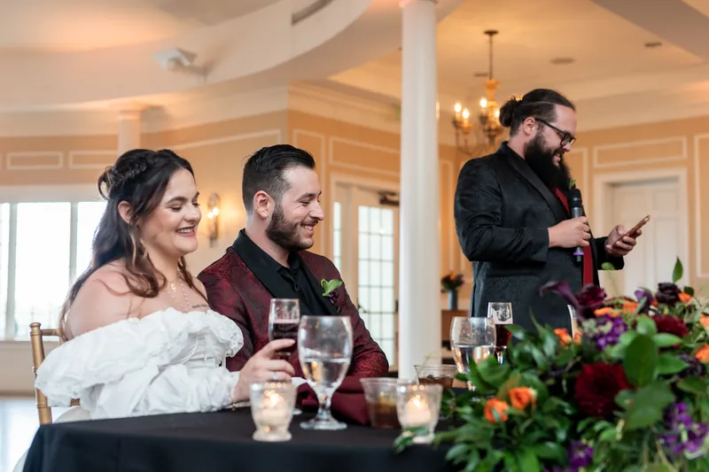 Alexandra and Jason sit together at their reception table as a speaker addresses the crowd at Riverhouse Events.