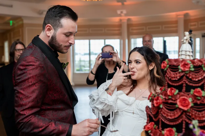 Jason and Alexandra feed each other cake beside towering red rose arrangements at their Riverhouse Events reception.