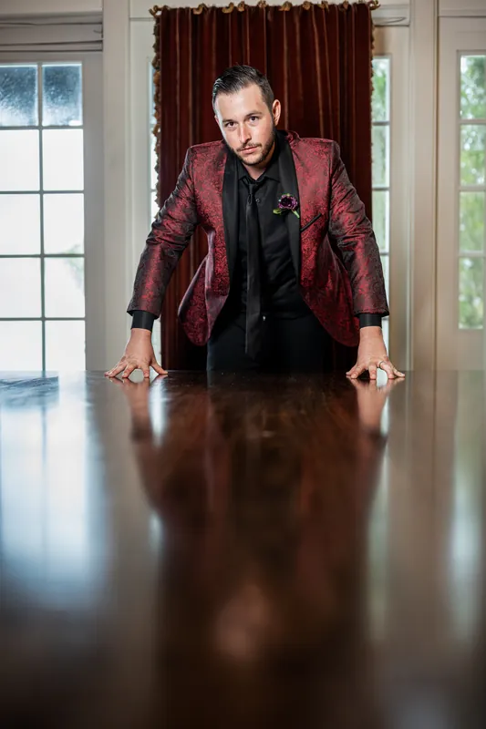 Jason leans forward on a polished table, wearing a burgundy jacket and black shirt at Riverhouse Events.