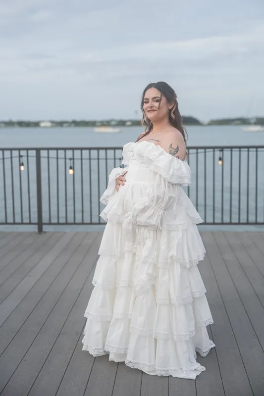 Alexandra stands alone on a waterfront deck in her layered white gown, smiling at the camera with water visible behind her.