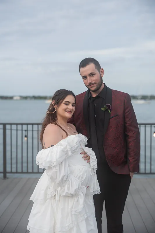 Alexandra and Jason stand on a riverside deck at Riverhouse Events, with Alexandra in a ruffled white off-shoulder gown and Jason in a burgundy blazer.