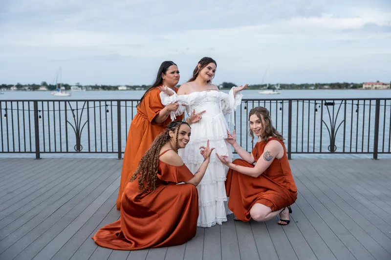 Three bridesmaids in terracotta robes adjust Alexandra's layered white dress on the waterfront deck at Riverhouse Events.