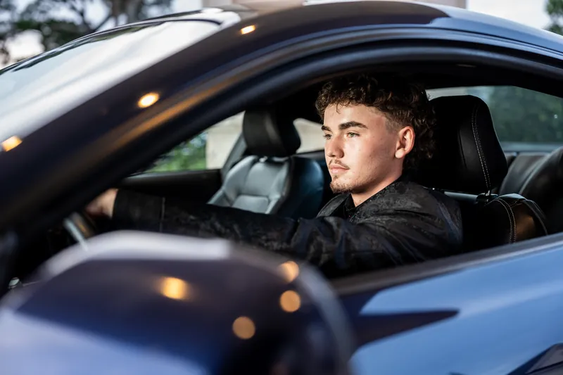 Jason sits behind the wheel of a blue car, looking out the window with a neutral expression.