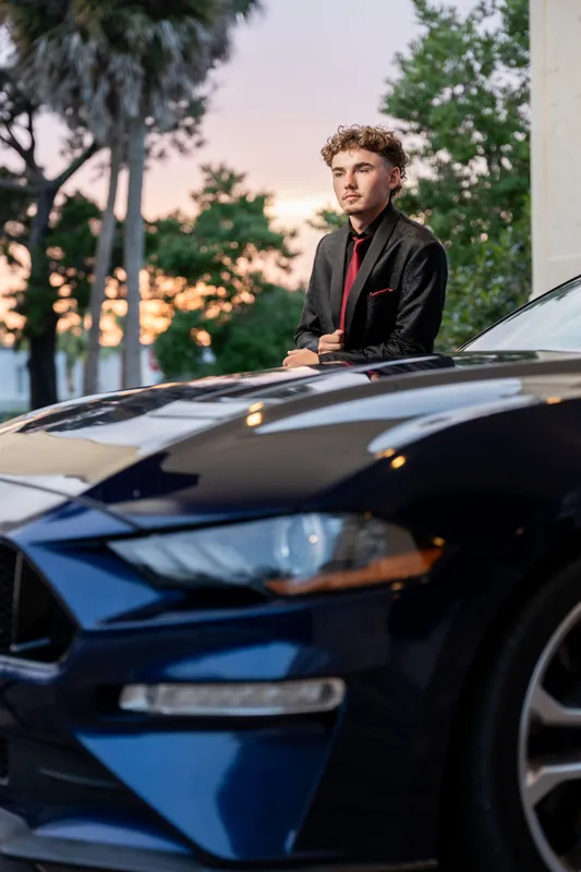 Jason leans against a blue car during dusk, wearing a burgundy shirt over a dark base layer.