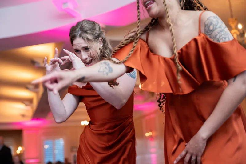 Women in rust-colored dresses laugh and dance together under pink uplighting at the reception.