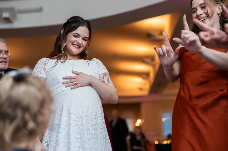 A woman in a white lace dress smiles while guests in orange dresses cheer nearby.