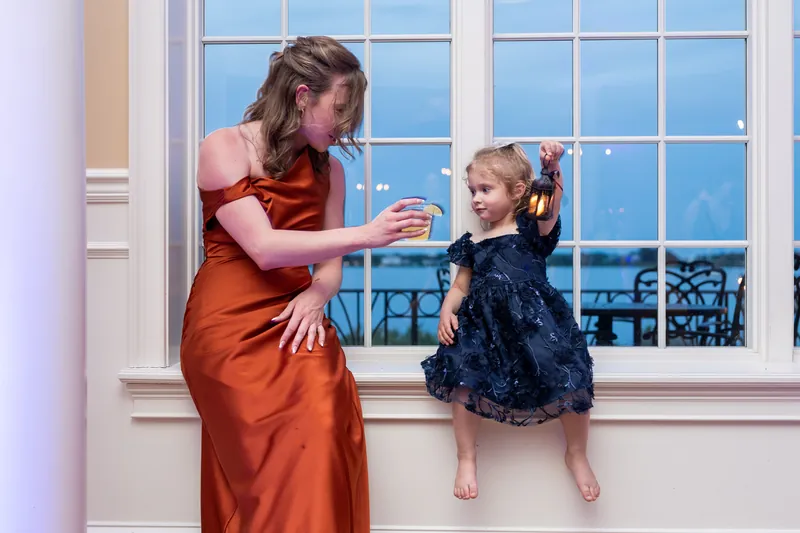 At Riverhouse Events, a woman in rust adjusts a young girl's navy dress by large windows overlooking water.