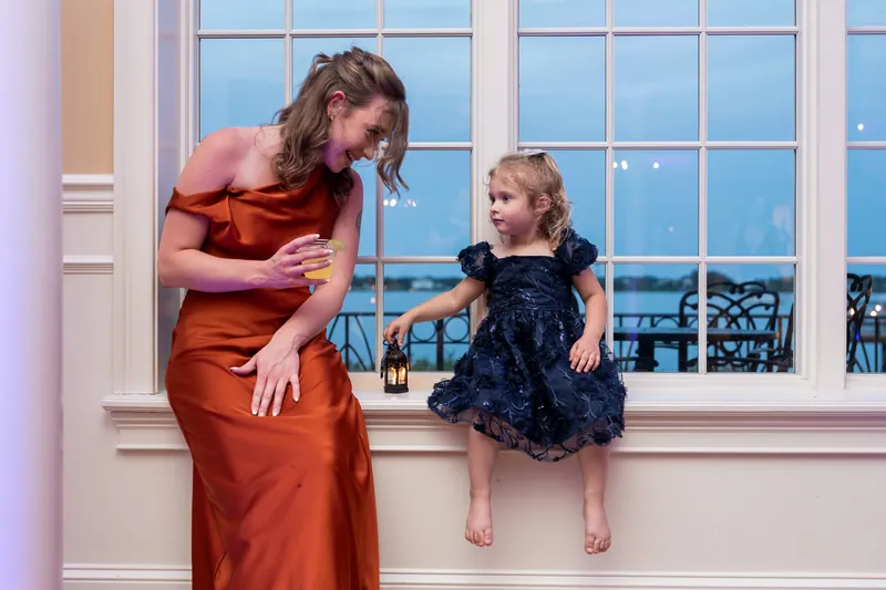 At Riverhouse Events, a woman in rust bends down to chat with a small girl in a navy dress by the windows.
