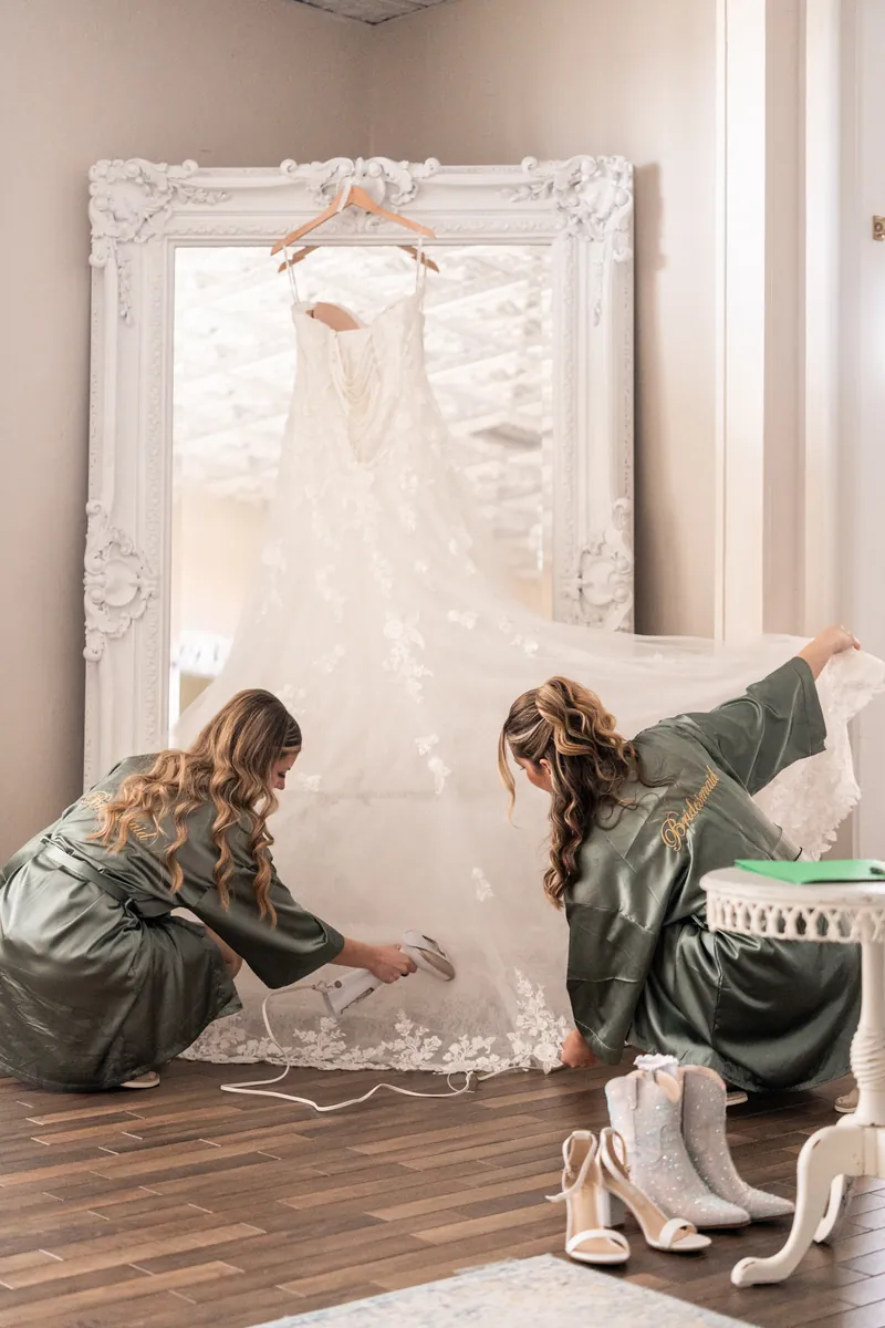 Bridesmaids in sage robes steaming wedding dress in front of ornate white mirror