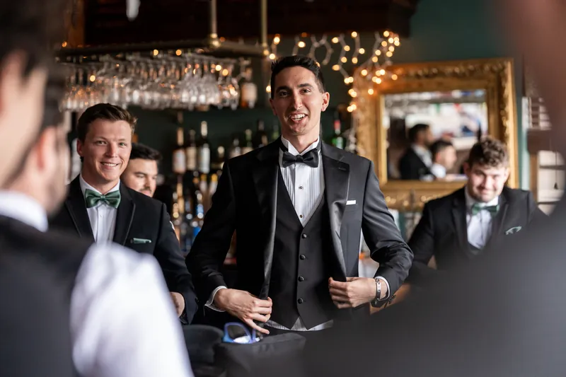 Groom laughing with groomsmen at bar with string lights and bottles
