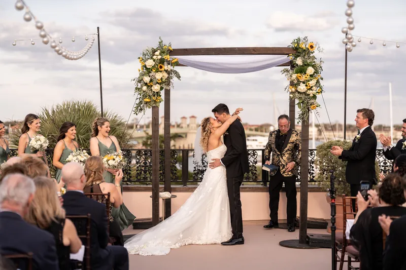 First kiss at rooftop ceremony with bridal party cheering and Matanzas Bay behind them