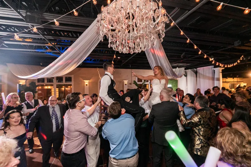 Hora with bride and groom lifted on chairs under crystal chandelier with glow sticks