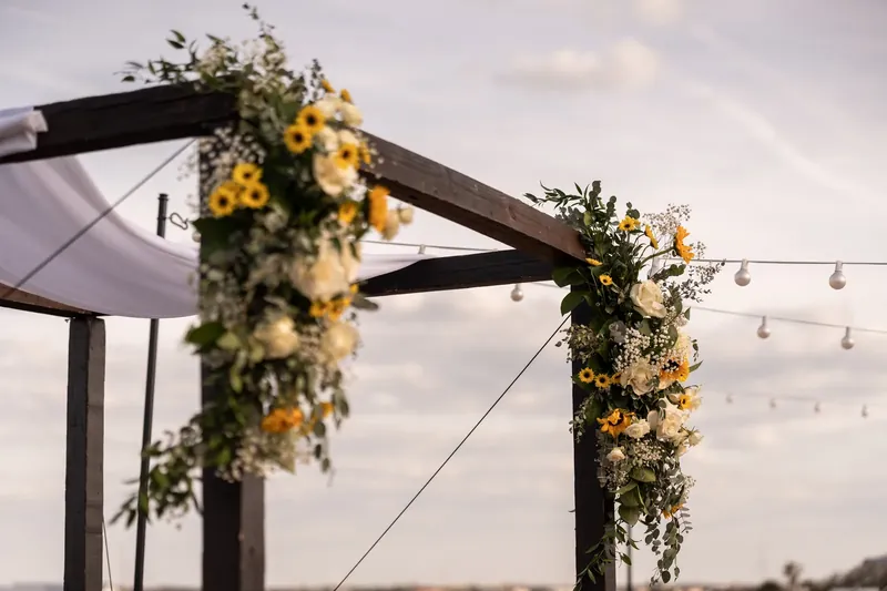Yellow blooms and cascading greenery drape from black pergola beams under an overcast sky at The White Room.