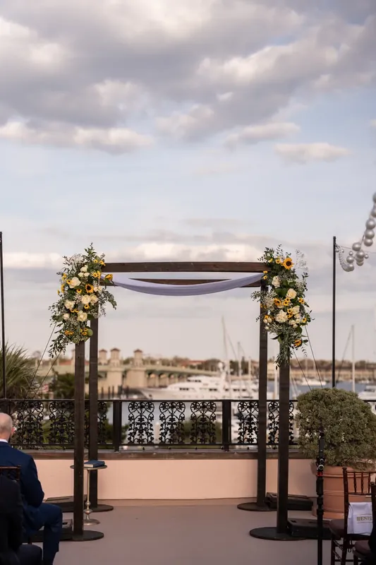 A black pergola with yellow and white floral garlands frames a view of St. Augustine's waterfront and historic buildings.