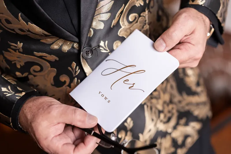 Hands holding a white card with "Alex & Dino" written in script, worn over a patterned jacket.