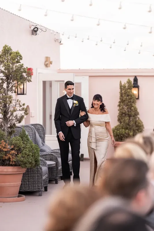 Dino in a black tuxedo and Alexis in a white off-shoulder gown walk together at The White Room's pink-hued venue.