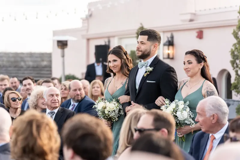 Dino and Alexis stand before seated guests during their ceremony, flanked by bridesmaids holding white bouquets.