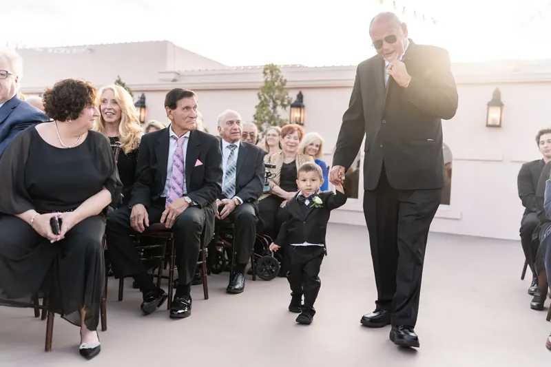 An older gentleman gestures while speaking to a young child during the outdoor ceremony as family members watch.