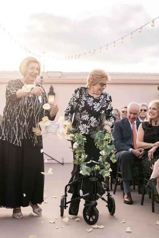 Guests smile as two women guide a flower-adorned stroller down the aisle during the processional.