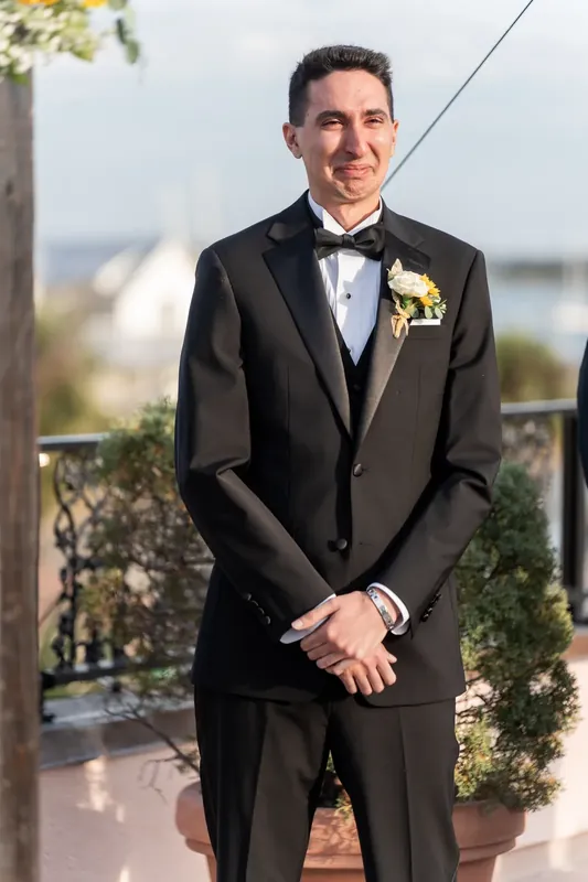 Dino smiles in his black tuxedo with boutonniere, positioned before the ceremony backdrop overlooking the city.