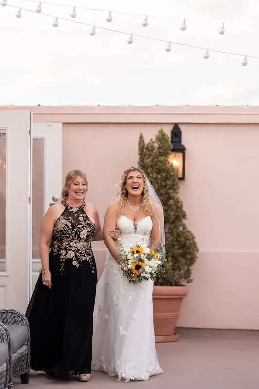 Alexis stands beside a woman in black on a pink terrace, holding her white bouquet before the wedding begins.