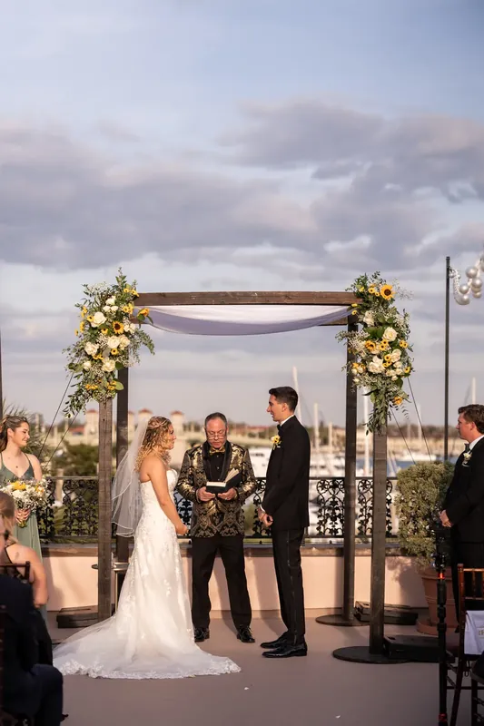 Alexis and Dino exchange vows beneath the decorated pergola with the officiant and wedding guests surrounding them.