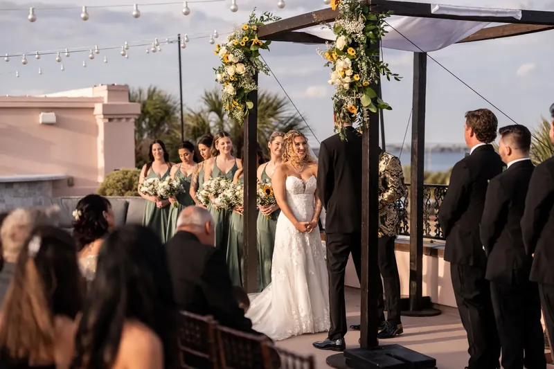 Alexis in her white wedding gown stands beneath a floral-adorned pergola as guests gather for the ceremony at The White Room.