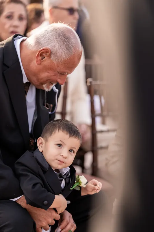 An older gentleman in a dark suit bends down to comfort a young boy in a matching suit during the wedding service.
