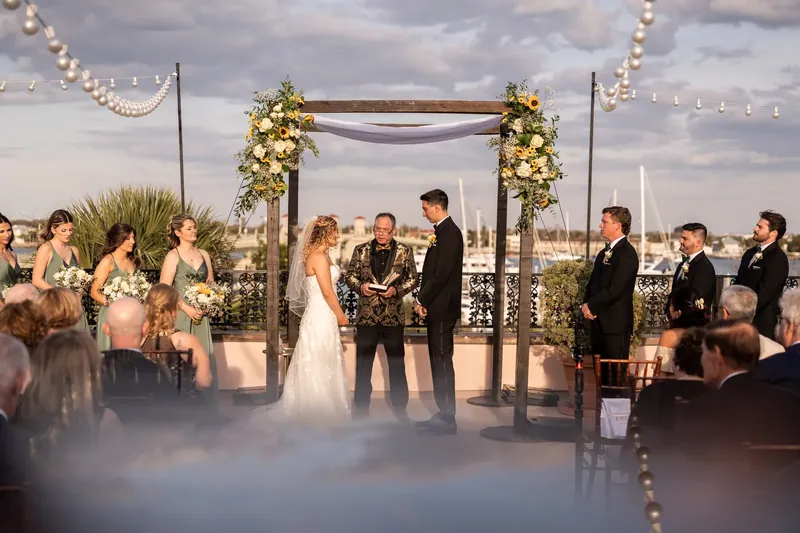 Dino and Alexis exchange vows beneath a decorated pergola with strung lights and greenery while guests watch nearby.