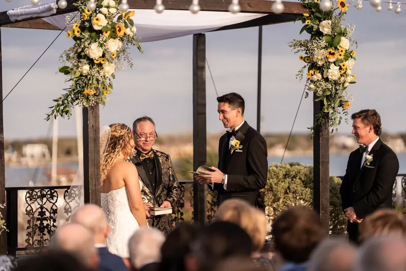 Alexis and Dino smile at each other during their vows as the officiant stands nearby in the candlelit pergola space.