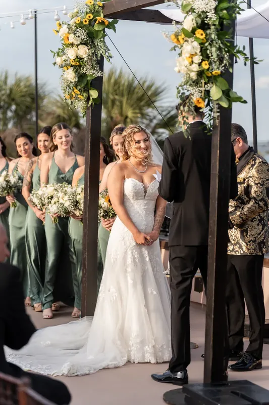 Alexis stands beneath a flower-draped pergola in her strapless lace gown as Dino and wedding party gather around them