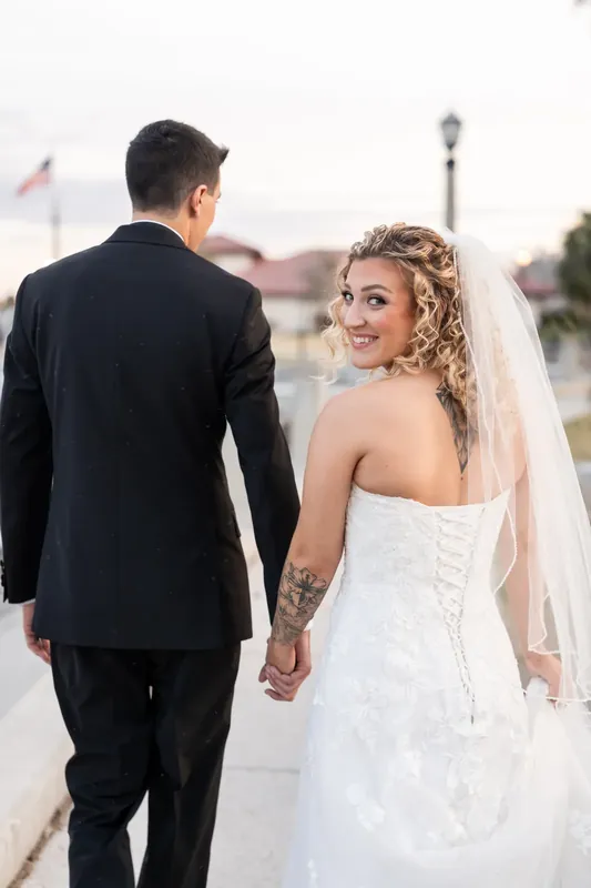 Alexis and Dino hold hands while walking along the waterfront, with Alexis looking back over her shoulder.