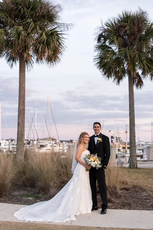 Alexis in an ivory gown and Dino in a black tuxedo stand together at St. Augustine Bayfront with sailboats and palm trees