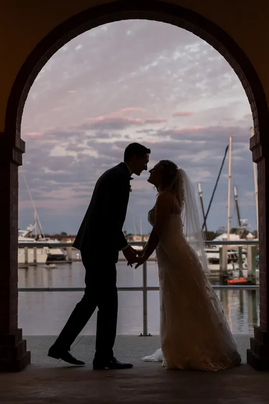 Dino leans in to kiss Alexis under a brick archway framing the water and moored sailboats at dusk.