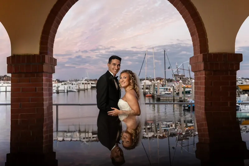 Alexis and Dino hold hands beneath a brick arch with their reflection mirrored in the wet ground, sailboats visible across