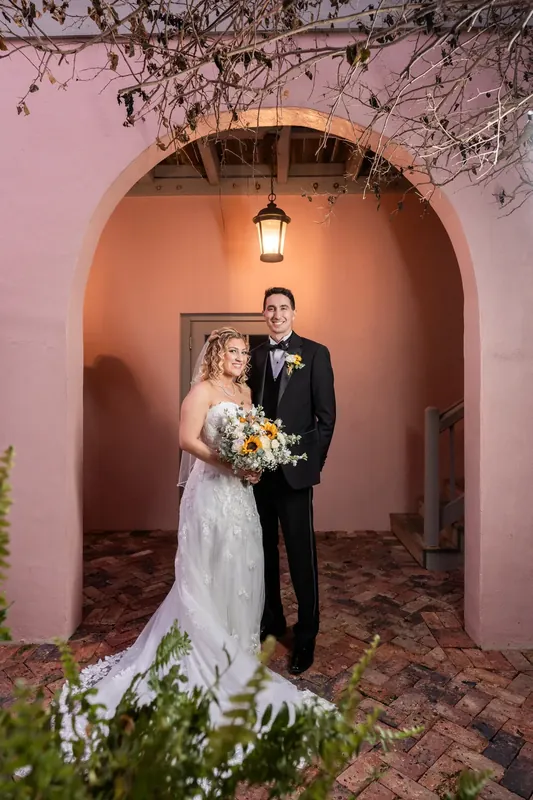 Alexis and Dino stand in a terracotta archway at The White Room, she holding white flowers and he in a black suit.